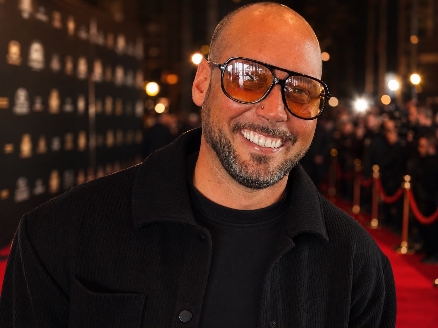 Ein lächelnder, bärtiger Mann mit getönter Sonnenbrille auf einem Event mit rotem Teppich.